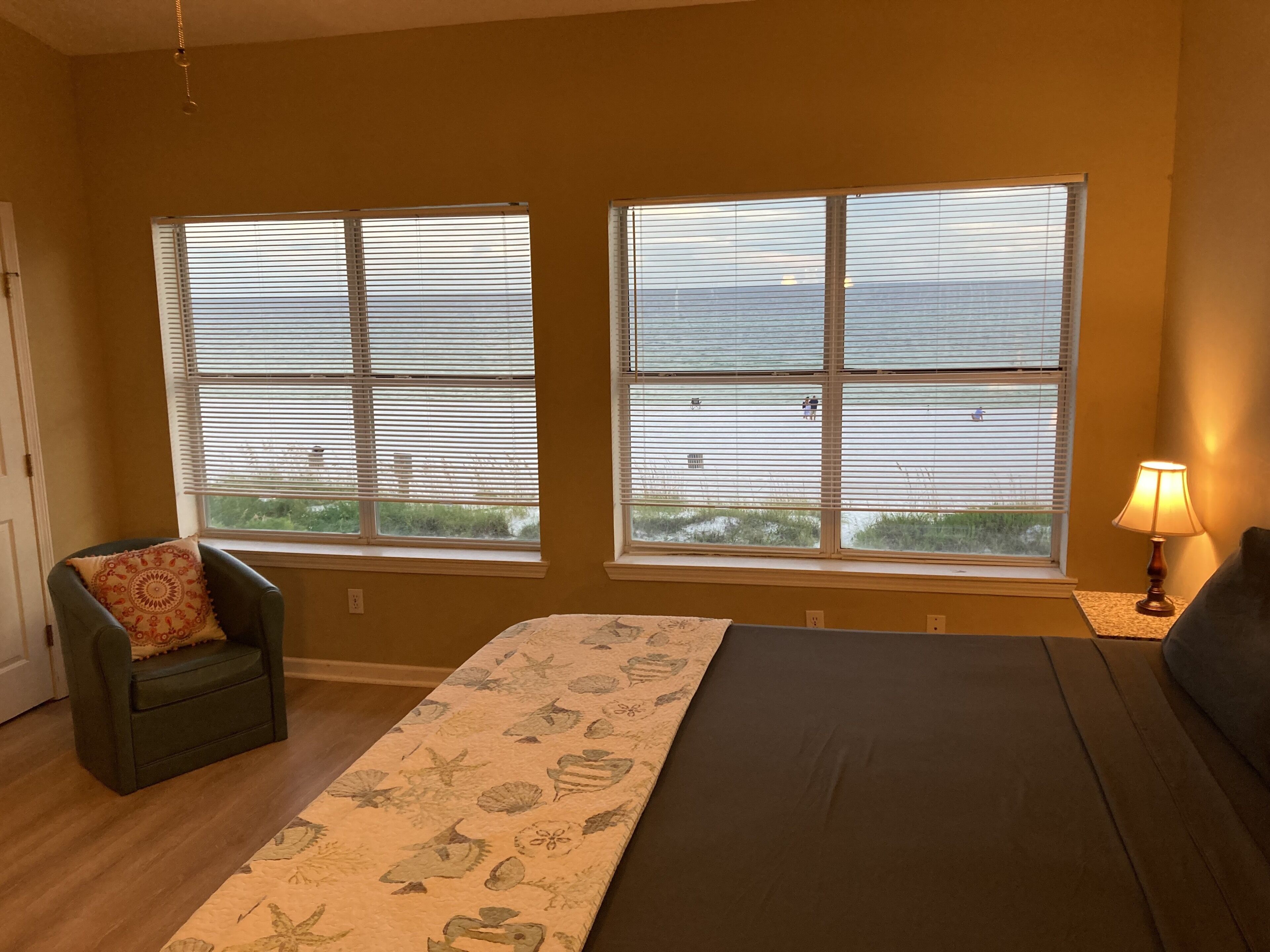 A bedroom with two large windows, a bed, a chair, and a lamp.