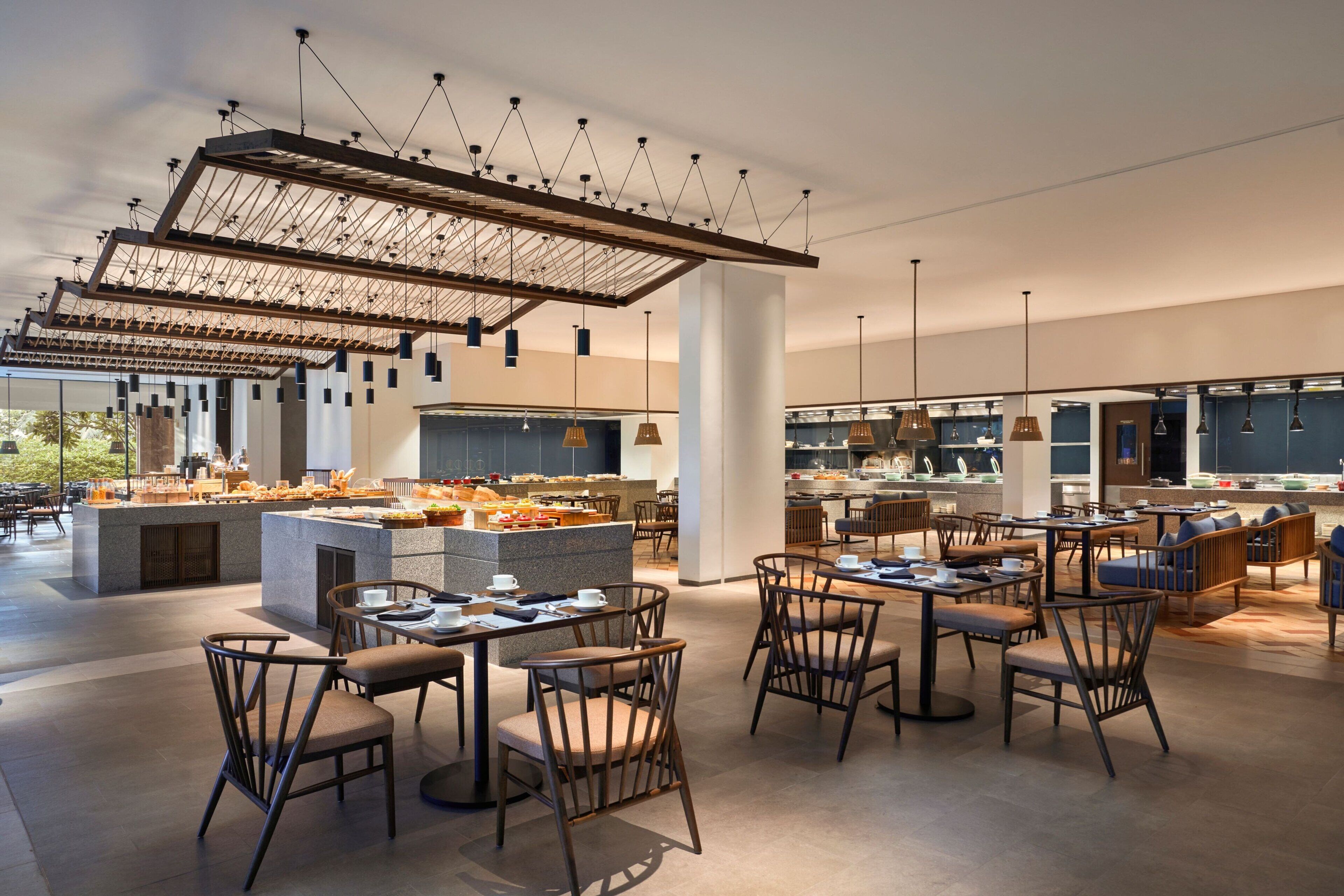 A modern cafe with multiple seating areas, a central counter with food display, and hanging pendant lights.