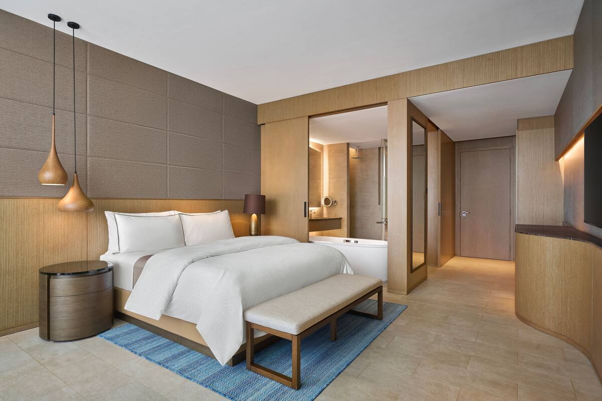 A modern hotel room with a large bed, bedside tables, a bench, and a bathroom visible through an open door.