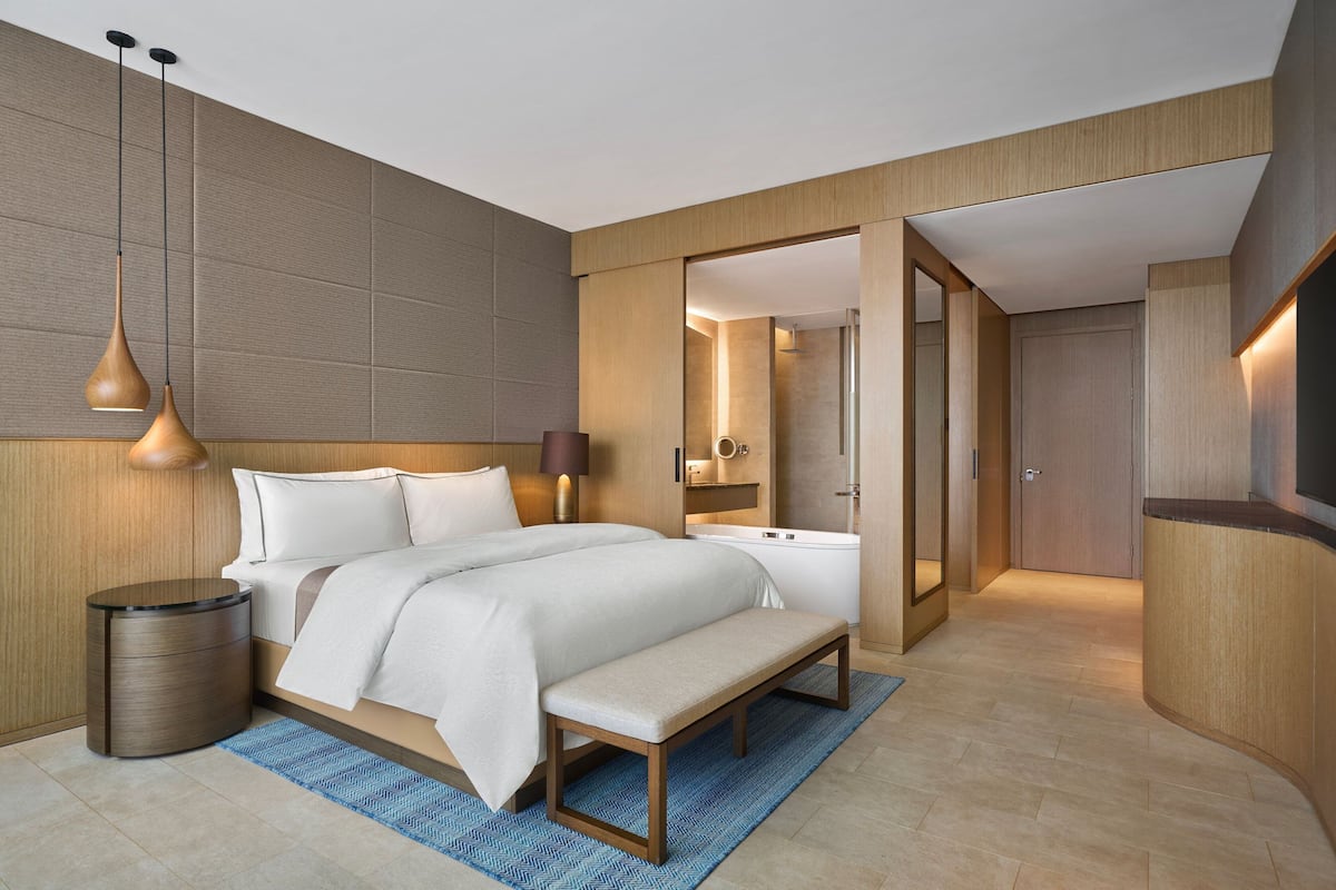 A modern hotel room with a large bed, bedside tables, a bench, and a bathroom visible through an open door.