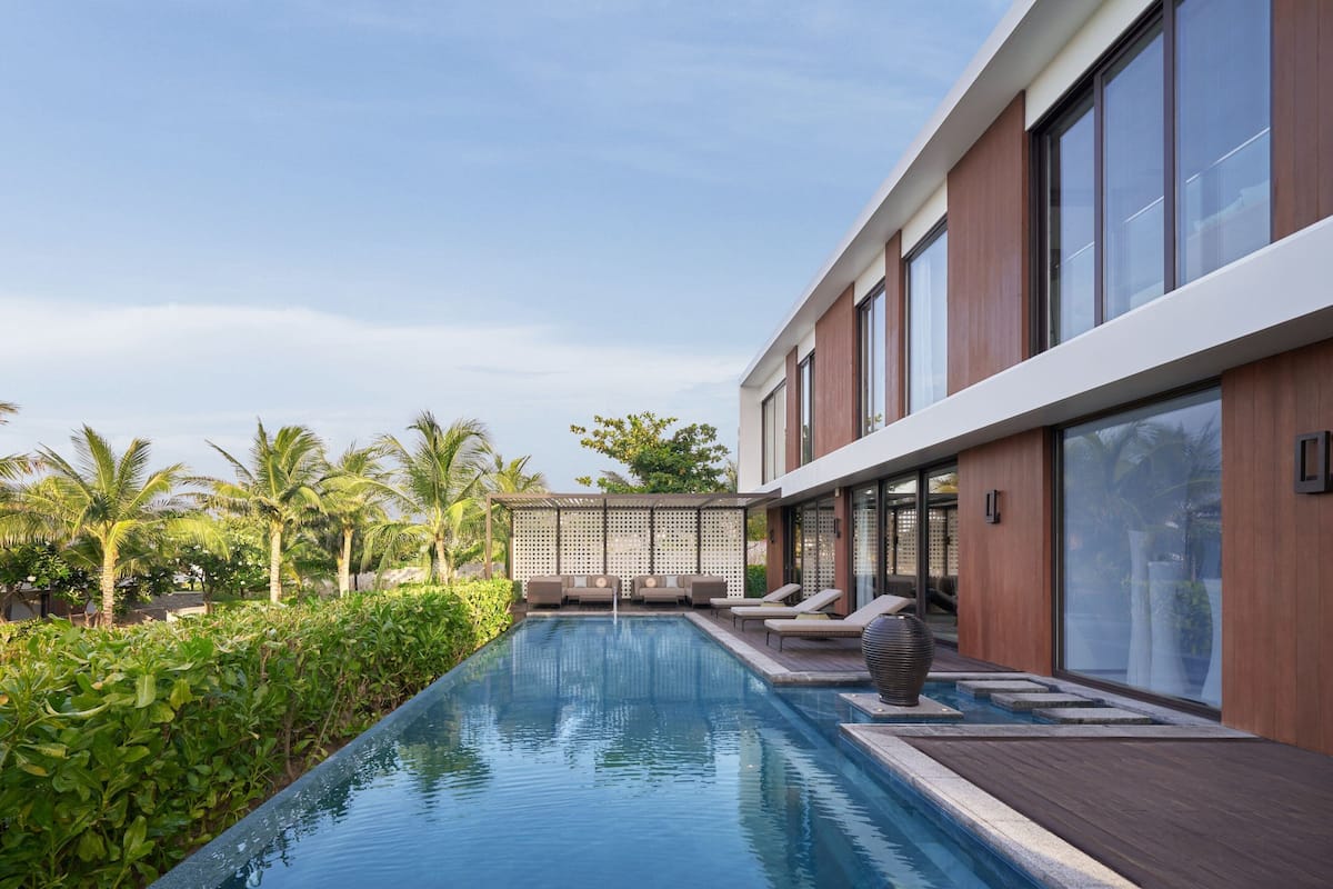 A modern pool area with a wooden deck, lounge chairs, and a view of the surrounding greenery and palm trees.