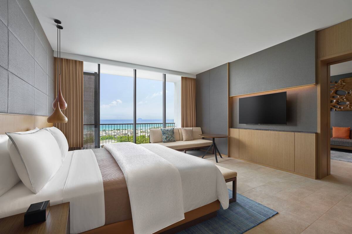 A modern hotel room with a large bed, a TV, and a balcony with a view of the sea.