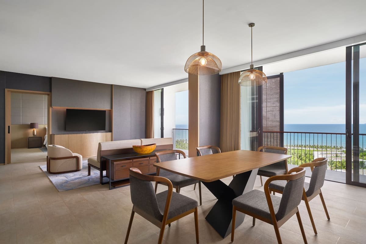 A modern living room with a wooden dining table, grey upholstered chairs, a large flat-screen TV, and a view of the ocean.