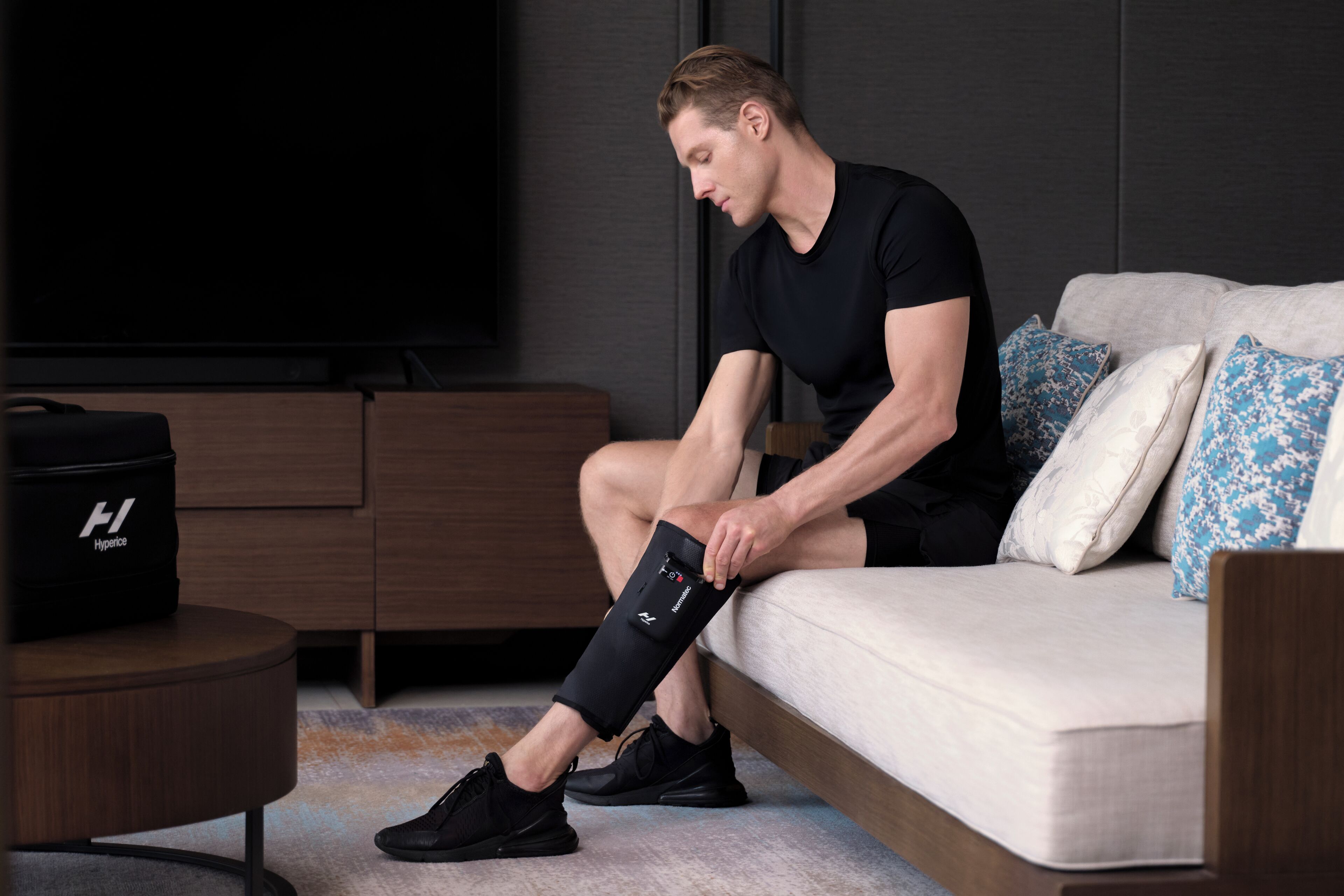 Una persona sentada en un sofá, con una ortesis de rodilla, cerca de un televisor y un bolso negro sobre una mesa de madera.