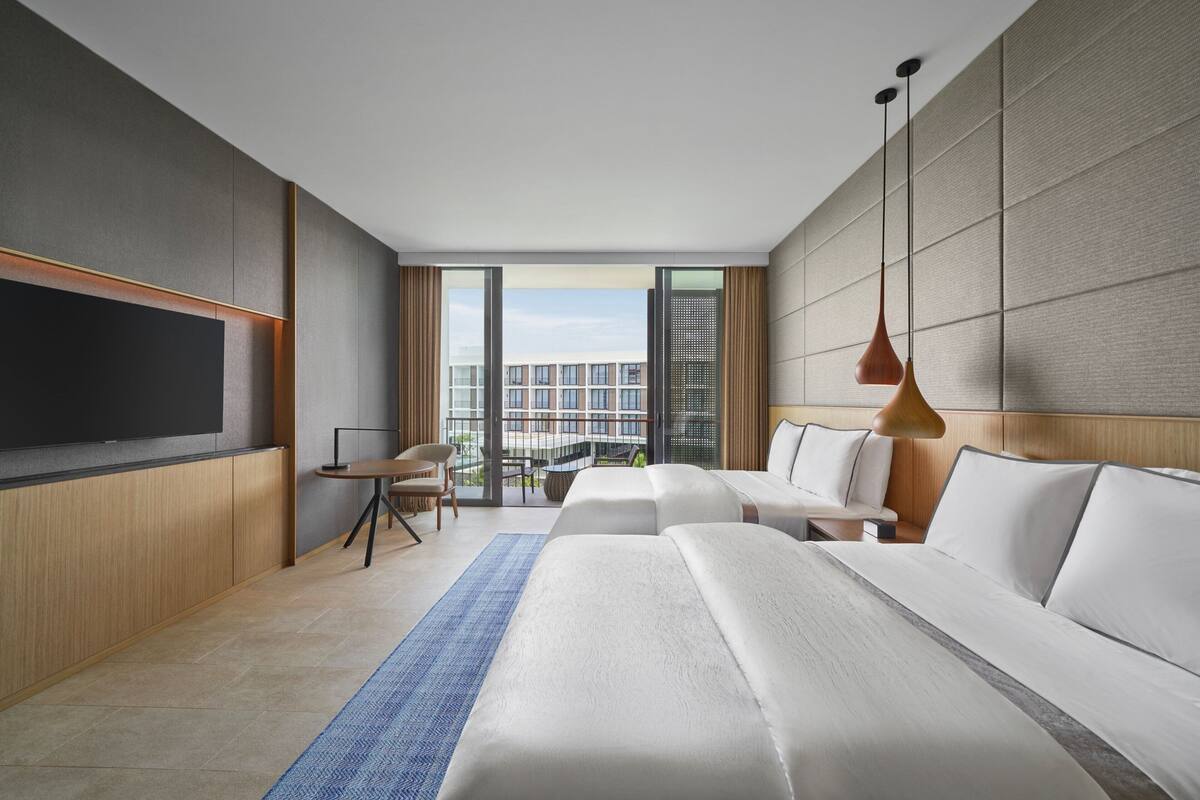 A modern hotel room with a large flat-screen TV, a sofa bed, a desk with a chair, and a view of the city through large windows.