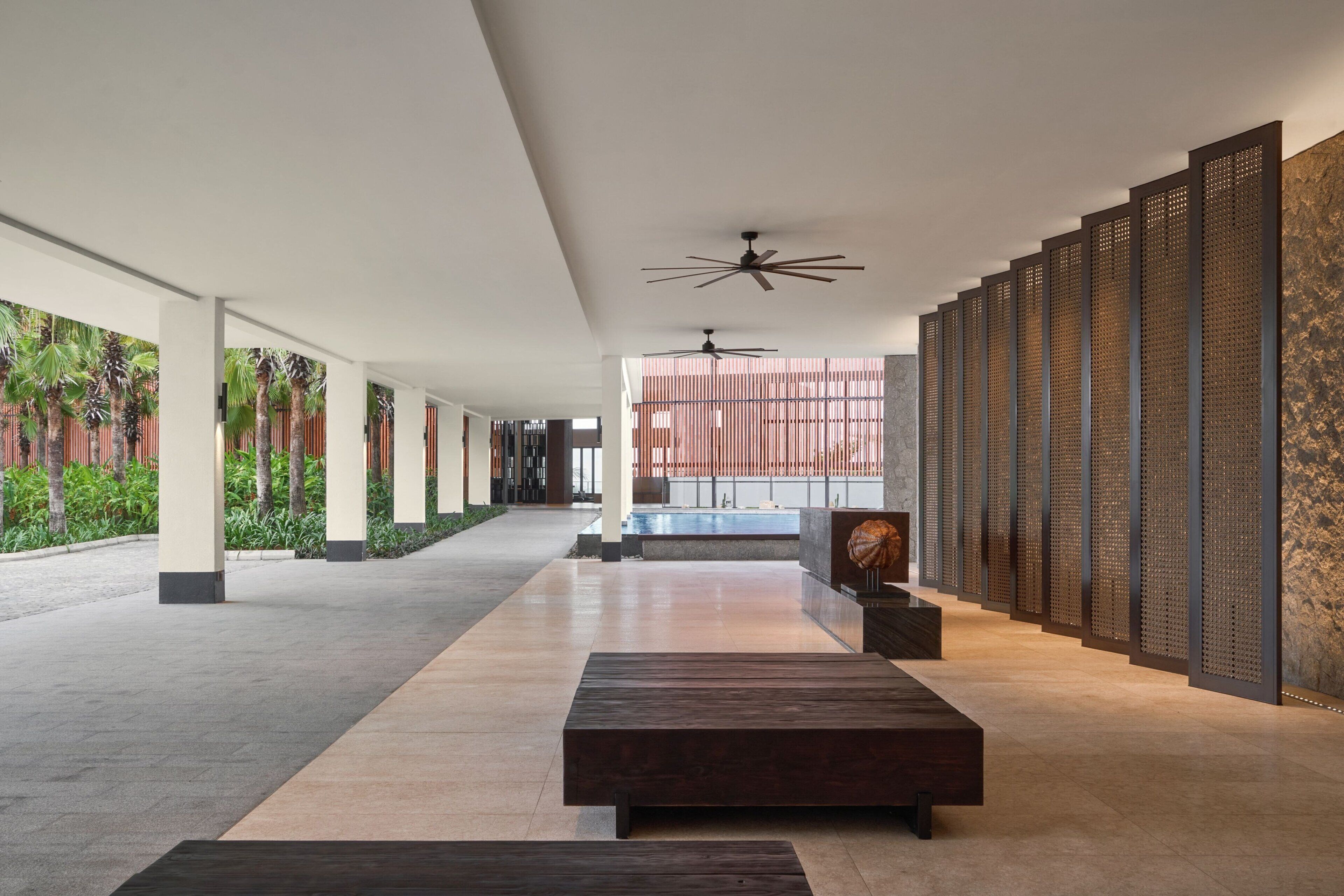 Un vaste couloir au design moderne, agrémenté d’un long banc, de ventilateurs de plafond et offrant une vue sur un espace piscine.