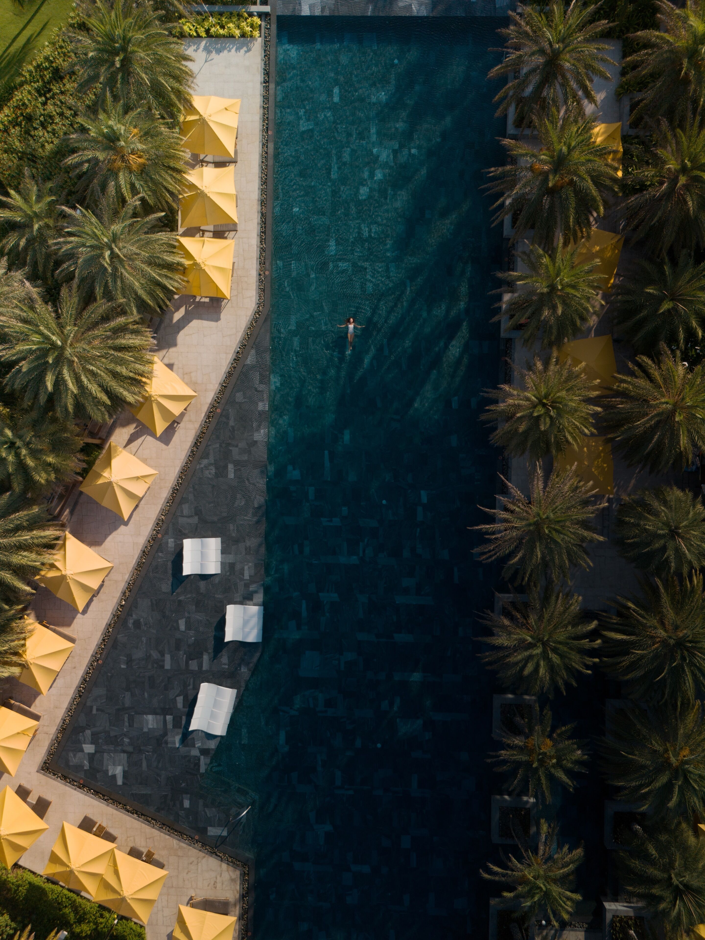 Luftaufnahme eines Schwimmbeckens mit Palmen und gelben Sonnenschirmen.