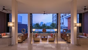 Un vestíbulo espacioso con un diseño moderno, que cuenta con una zona de estar central con sofás y sillones, una mesa de centro redonda y un amplio ventanal con vistas a palmeras y edificios.