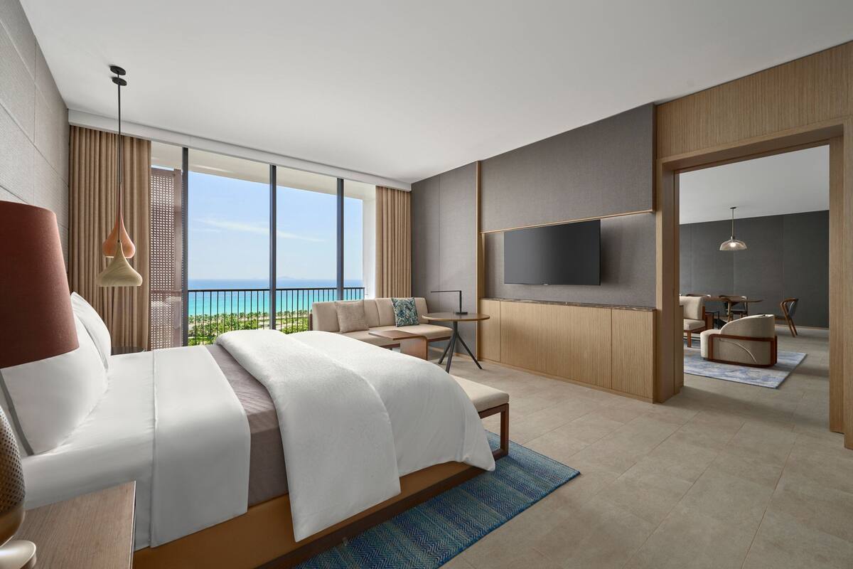 A modern hotel room with a large bed, a TV mounted on the wall, a balcony with a view, and a seating area with a table and chairs.