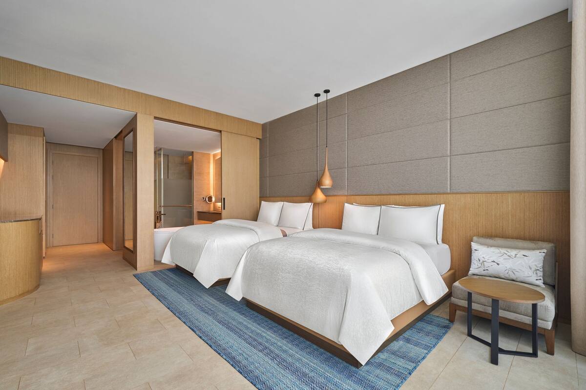 A modern hotel room with two beds, a small table, and a blue rug.