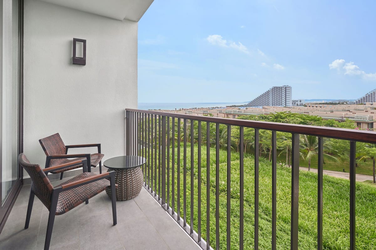 A balcony with two wicker chairs and a round table, overlooking a landscaped area with a view of distant buildings.