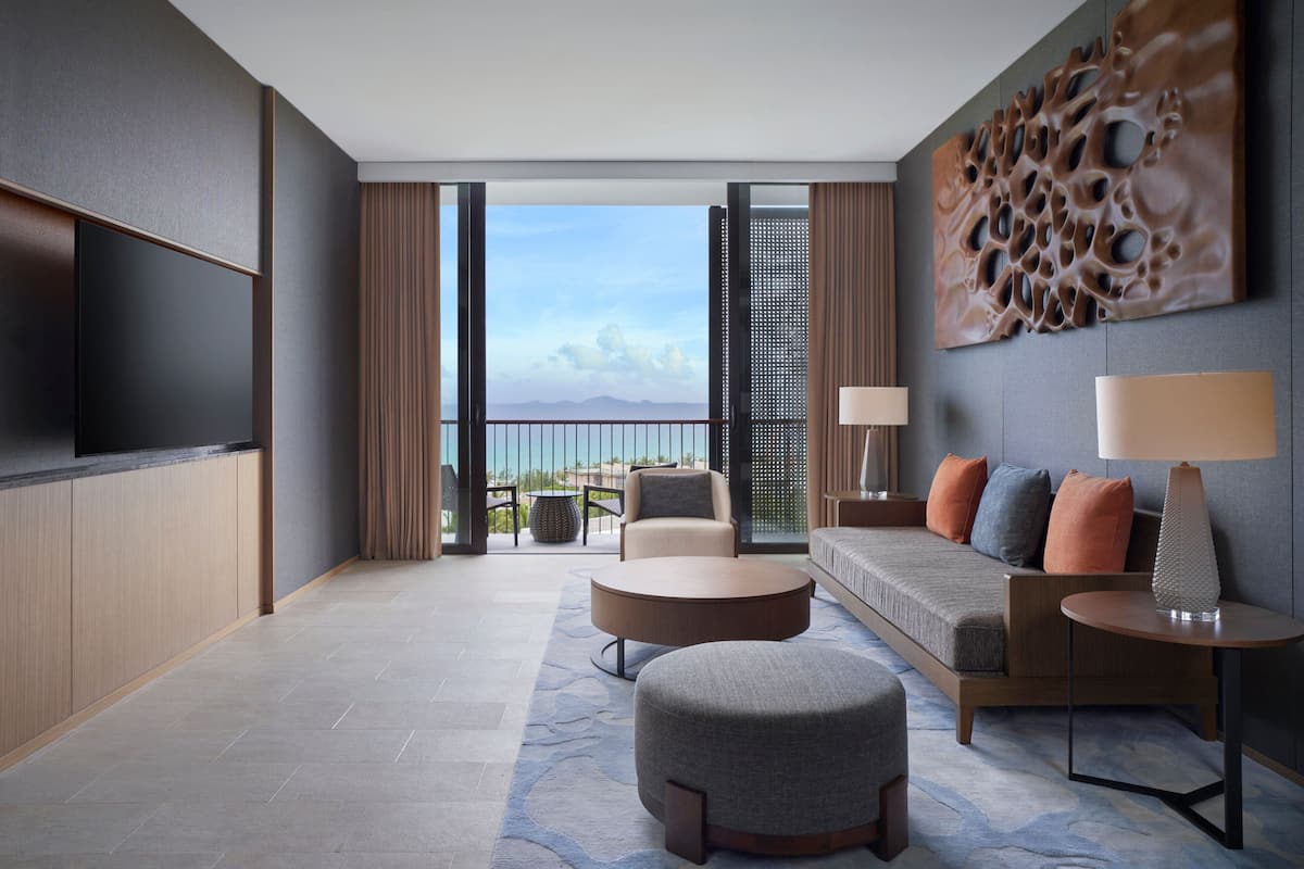 A modern hotel room with a large TV, a sofa, a round coffee table, and a balcony with a view of the city.