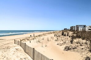 Ein Strand mit einer Holzlattenveranda, sandigen Dünen und einem mehrstöckigen Gebäude im Hintergrund.
