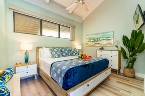 Quarto com cama, mesas de cabeceira, cômoda, janela grande com persianas, ventilador de teto, pintura do oceano e planta em vaso.