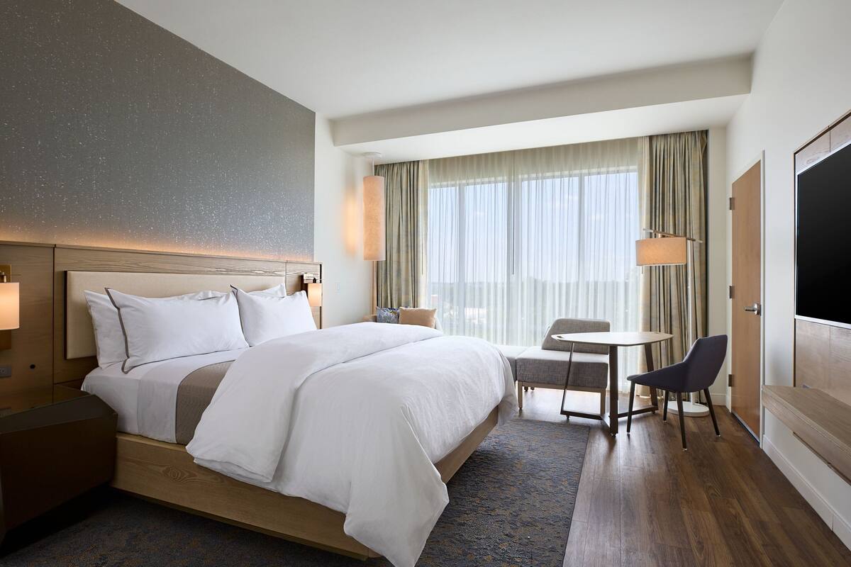 A modern hotel room with a large bed, bedside tables, a small desk with a chair, a large window with curtains, and a flat-screen TV.
