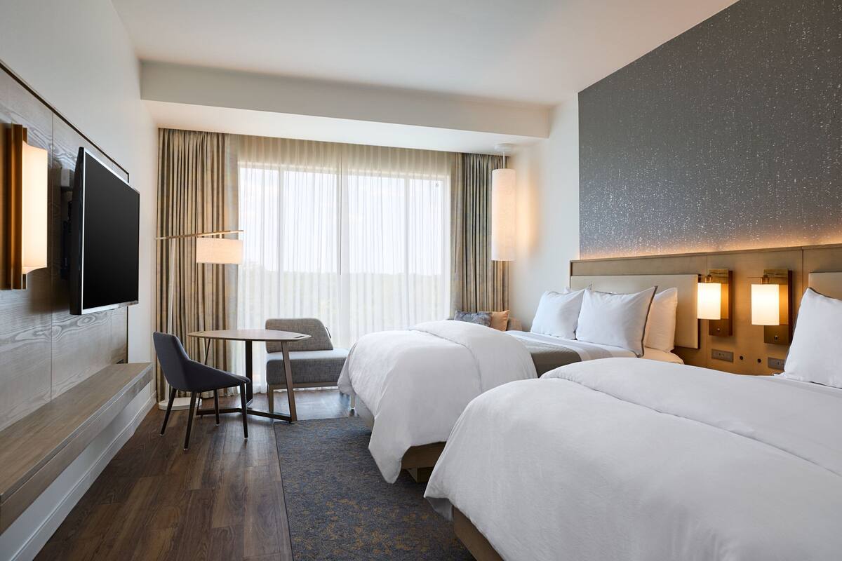 A hotel room with two beds, a television, a desk, and a large window with curtains.