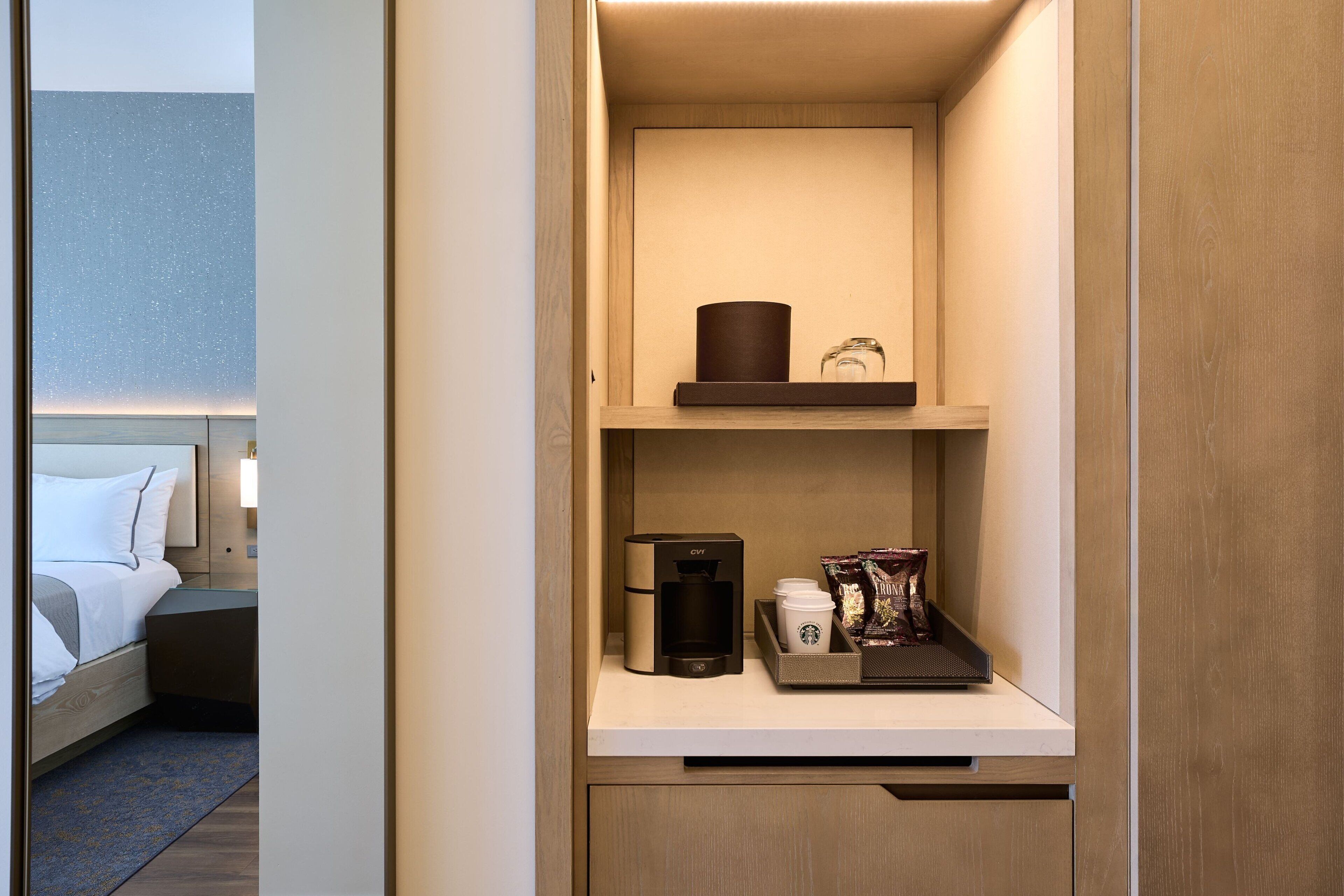 A hotel room with a neatly organized minibar, a bed with white bedding, and a nightstand with a lamp.
