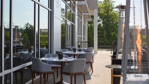 Un área de comedor al aire libre con mesas y sillas, ventanas amplias y vistas a la vegetación.