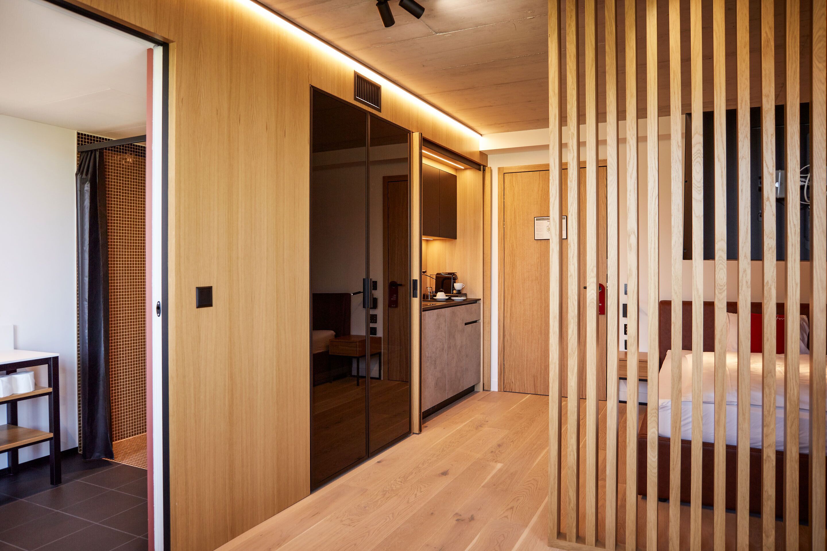 A modern hotel room with a wooden floor, a bed with white bedding, a small desk, and a sliding door leading to a bathroom.
