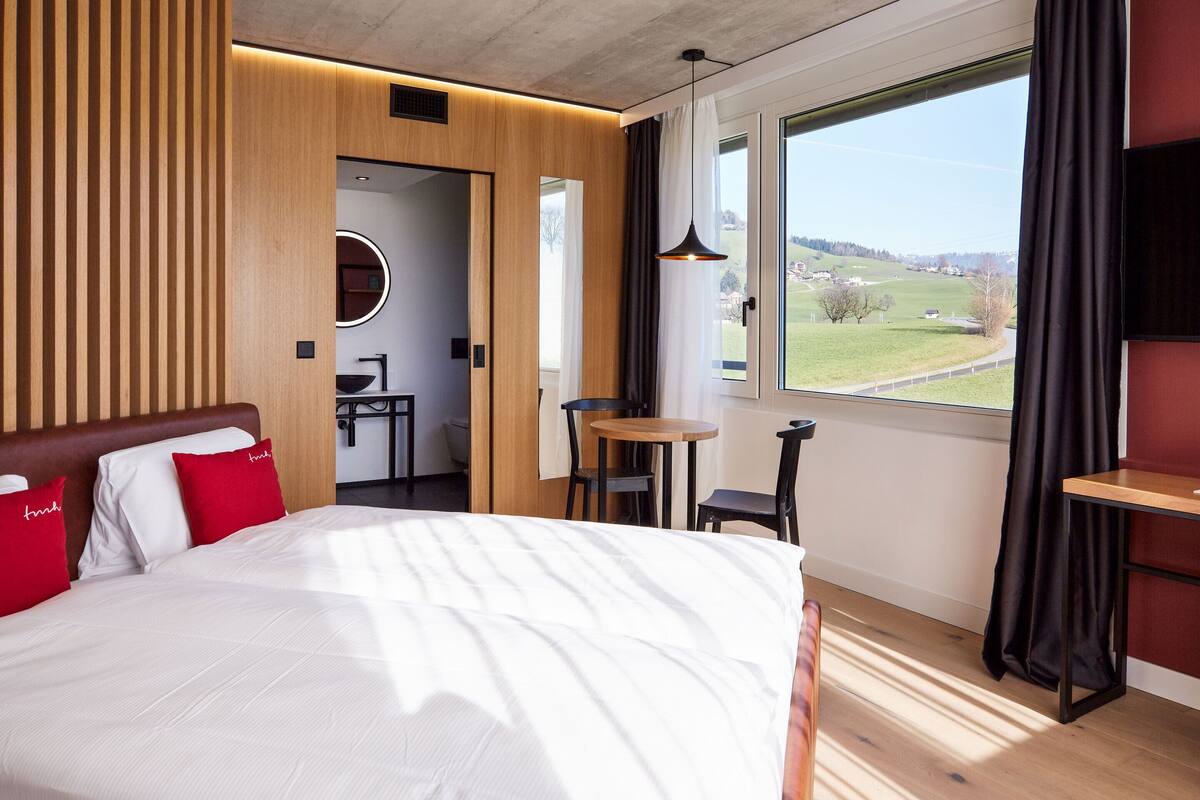 A modern hotel room with a large bed, a small table, a chair, a round mirror, and a view of the countryside.