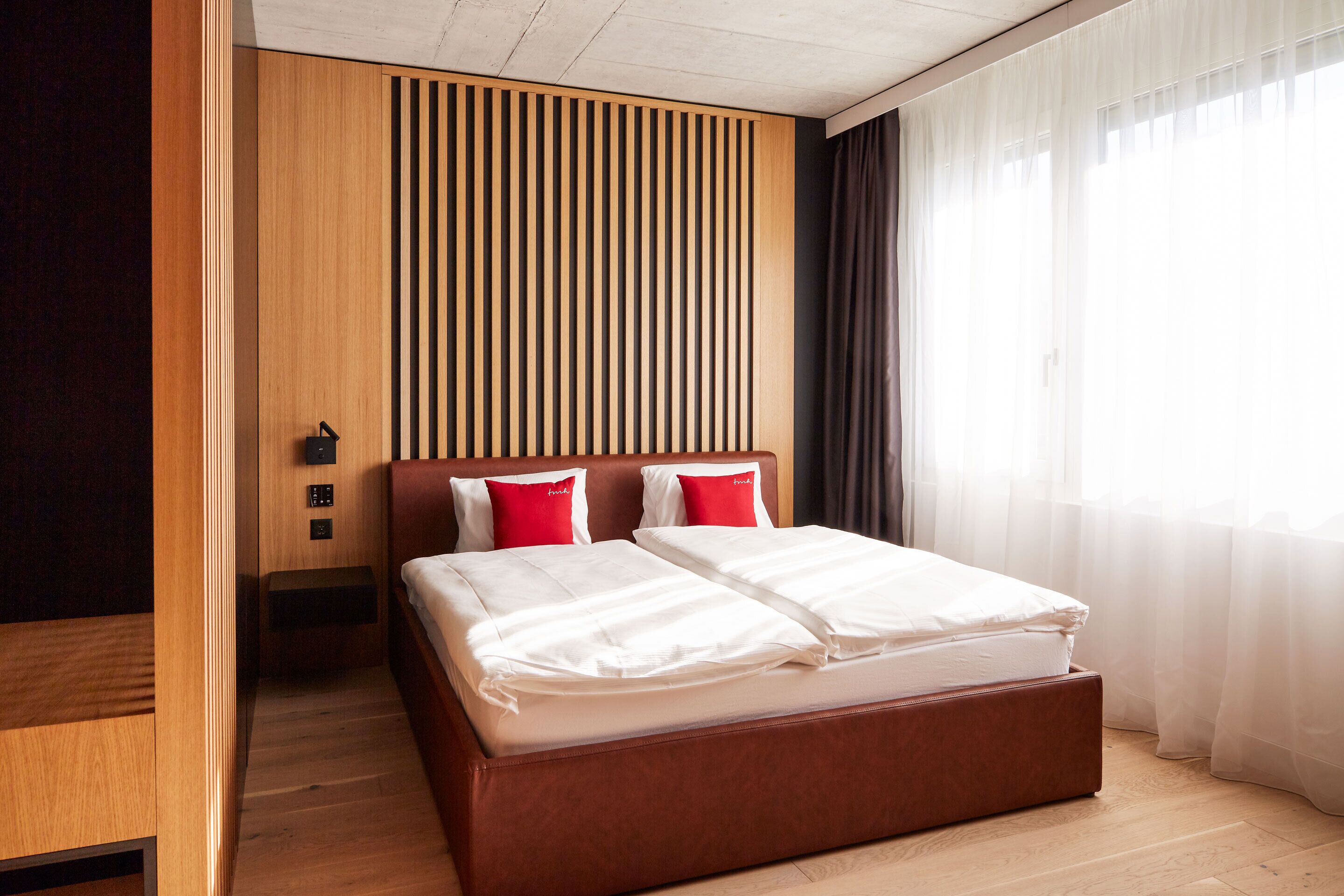 A modern hotel room with a wooden headboard, a large bed with white bedding, and a view of a cityscape through sheer curtains.