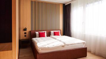 A modern hotel room with a wooden headboard, a large bed with white bedding, and a view of a cityscape through sheer curtains.