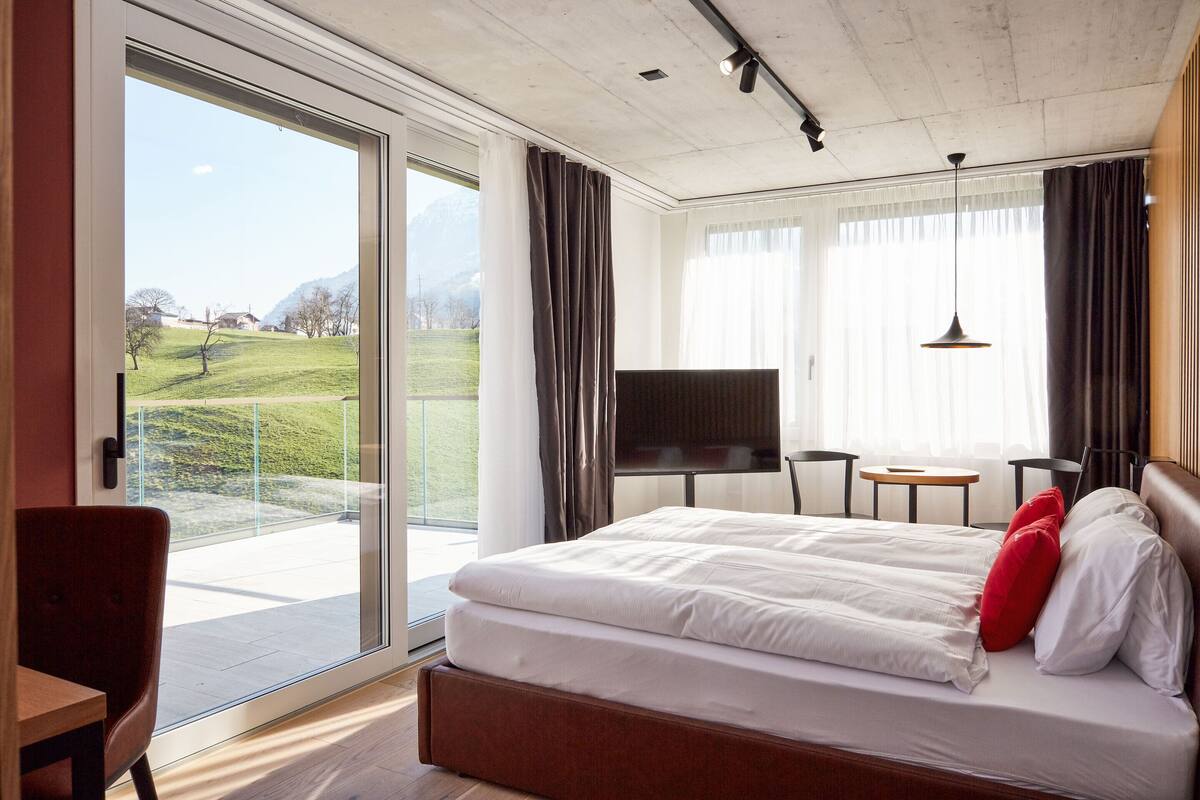 A hotel room with a large bed, a TV, a small table, and a view of a green landscape.