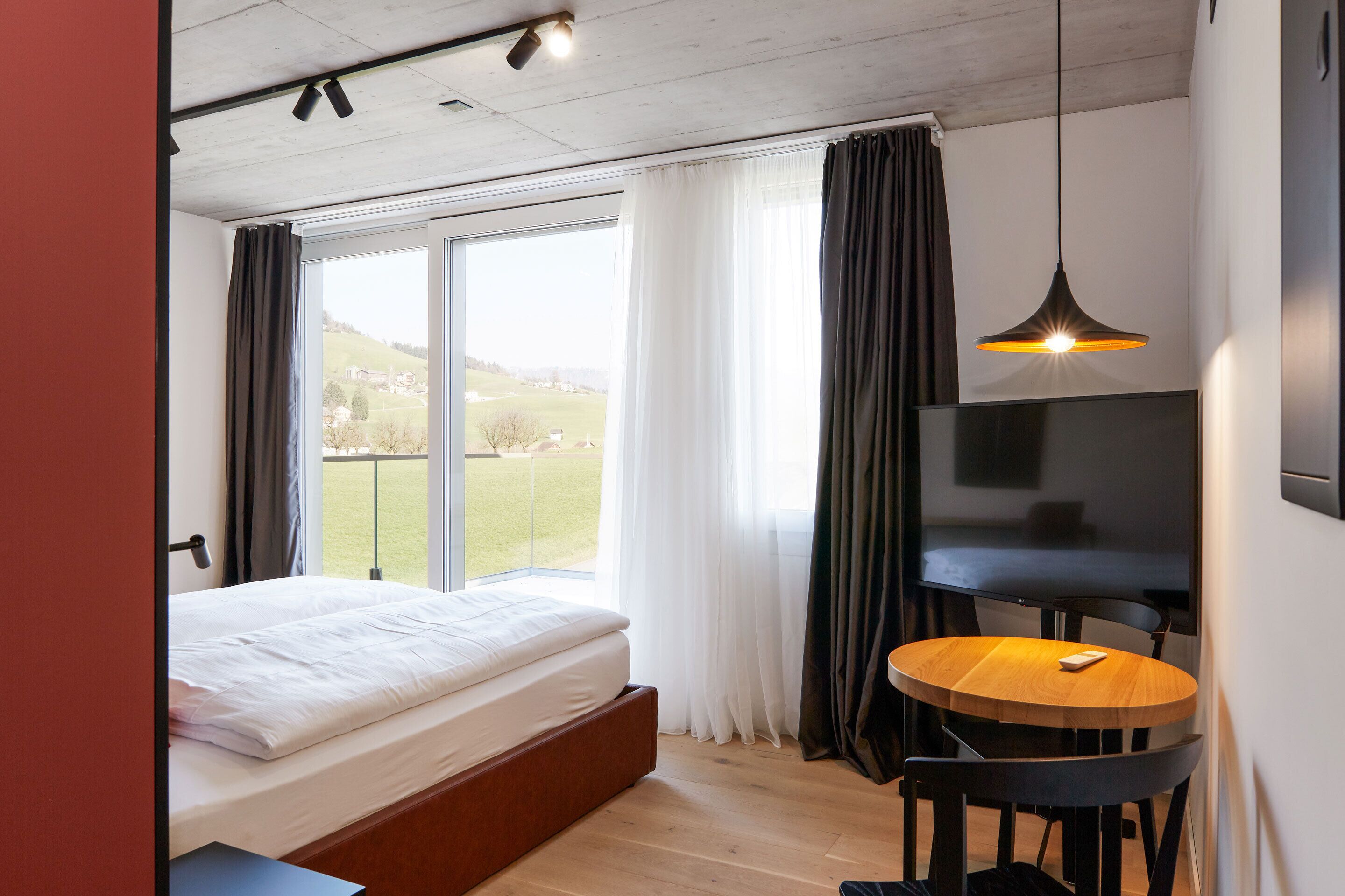 Un dormitorio moderno con un ventanal grande, una cama, un televisor y una pequeña mesa de madera.