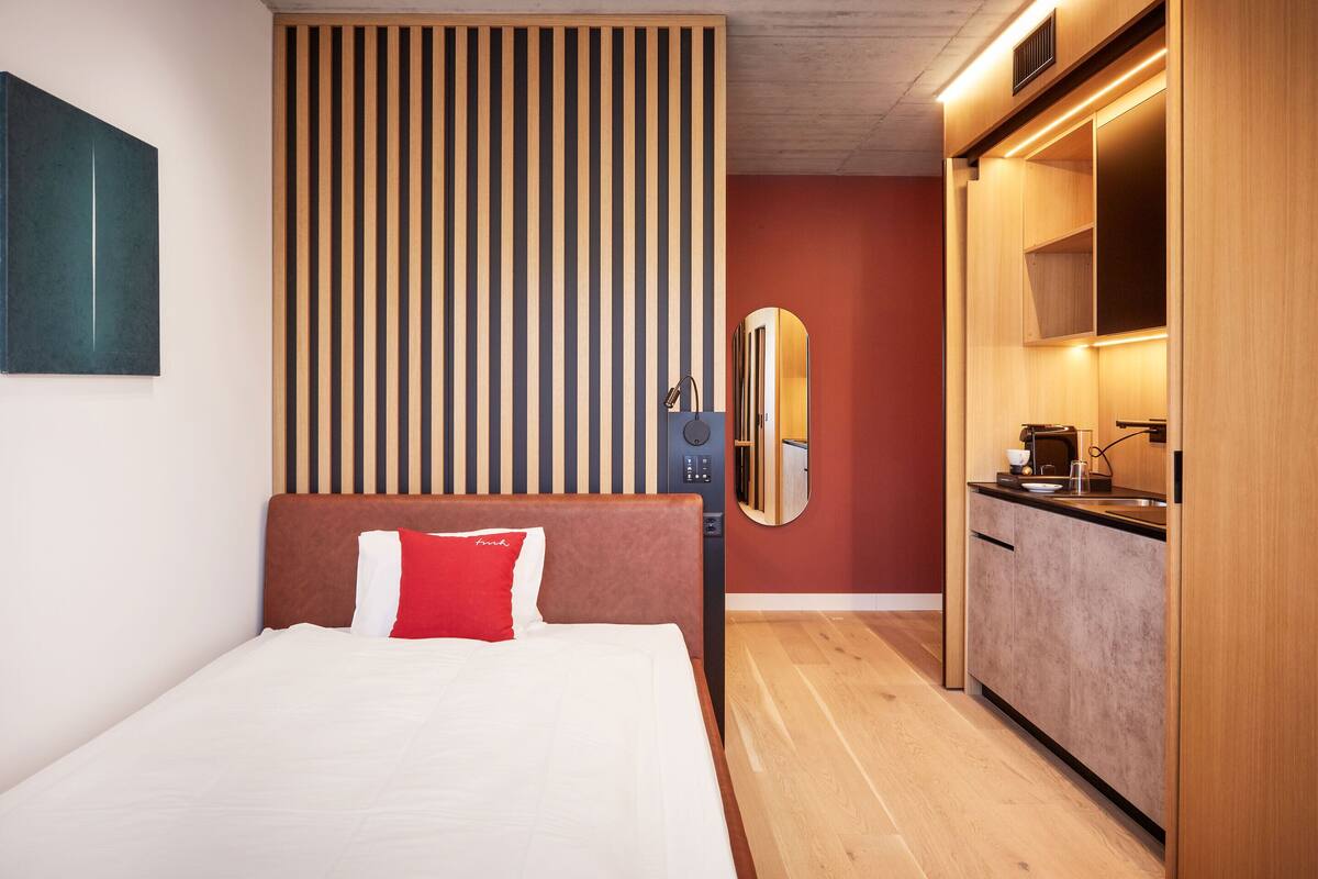 A hotel room with a bed, a wooden headboard, a vertical striped panel, a mirror, and a kitchenette.