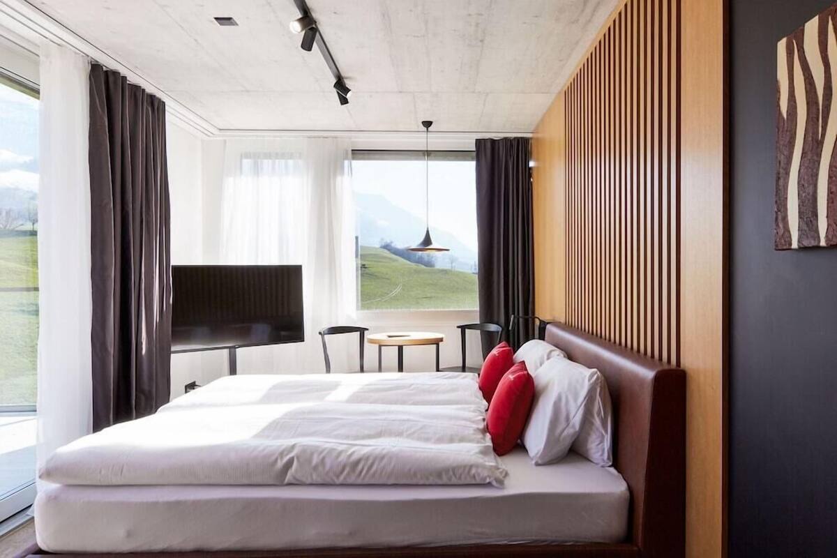 A modern hotel room with a large bed, a TV, a small table, and a view of the outdoors.