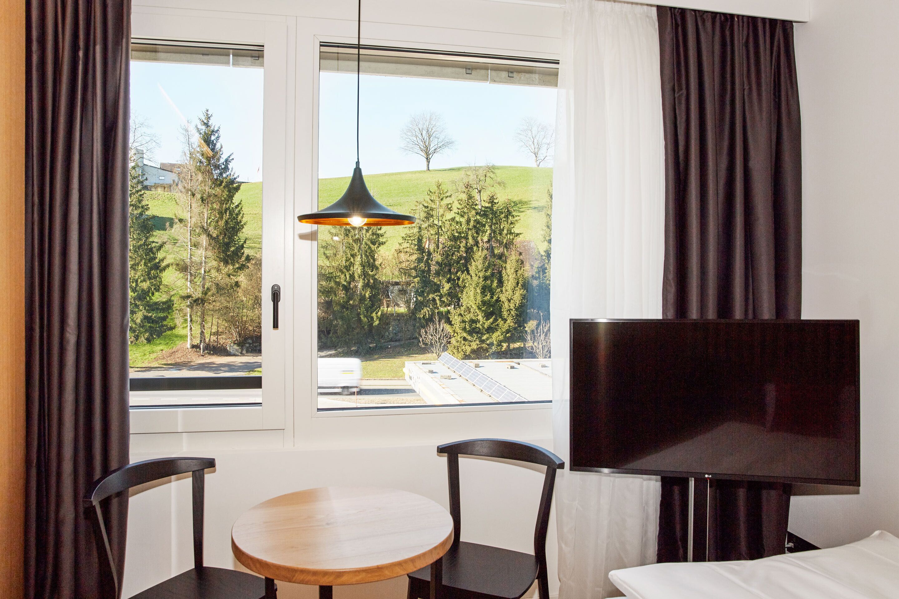 A room with a large window, a round table, two chairs, a TV, and a bed.