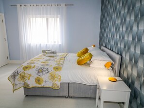 A modern bedroom with a large bed, a bedside table with a lamp, and a window with sheer curtains. - Wee Andy's (Buncrana)
