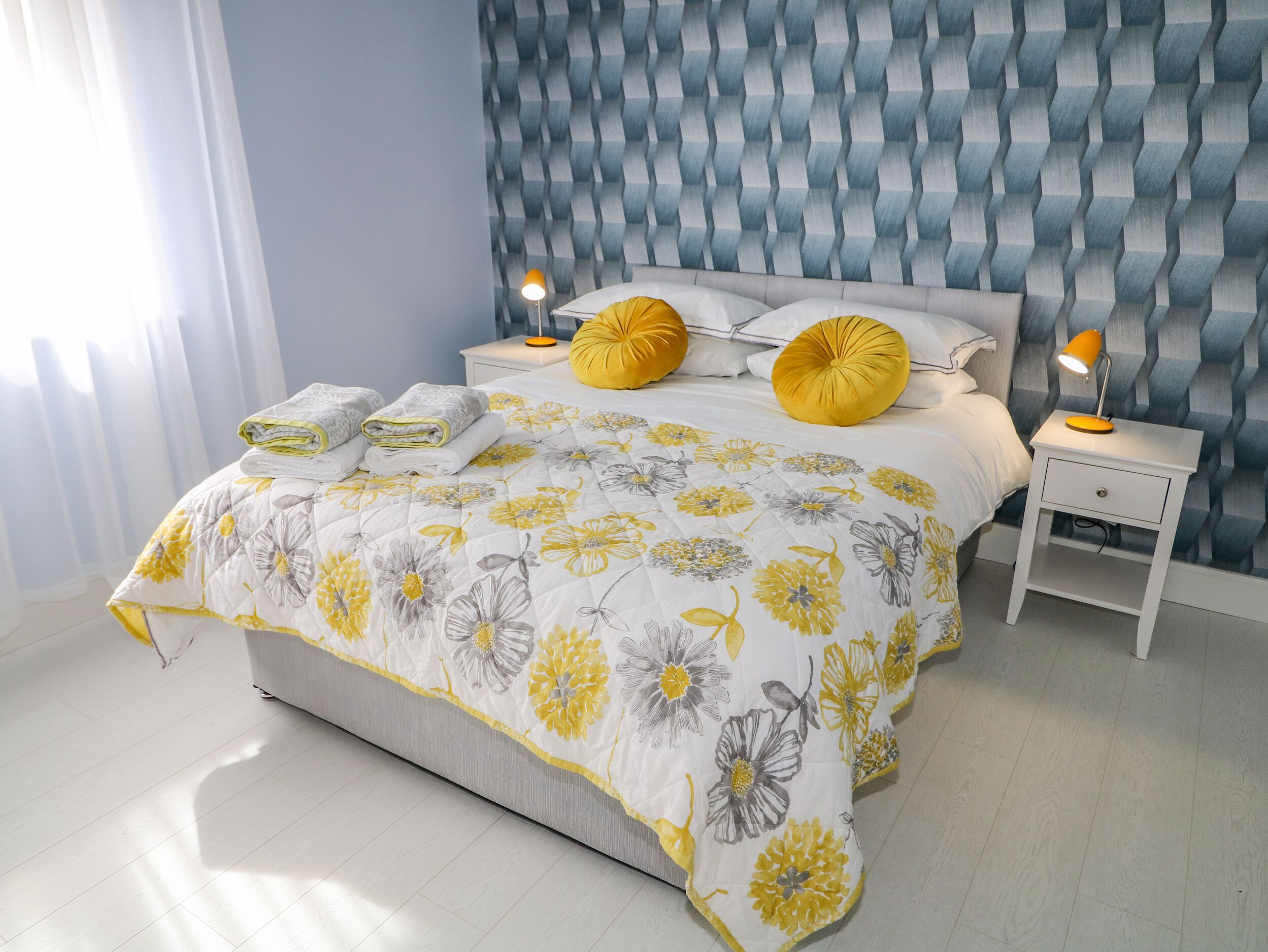 Una cama bien tendida con un edredón floral, dos almohadas amarillas y una lámpara de noche sobre una mesita blanca.