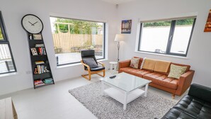 A modern living room with a leather sofa, a glass coffee table, a black leather armchair, a bookshelf, and a large window with a view of the outdoors. - Wee Andy's (Buncrana)