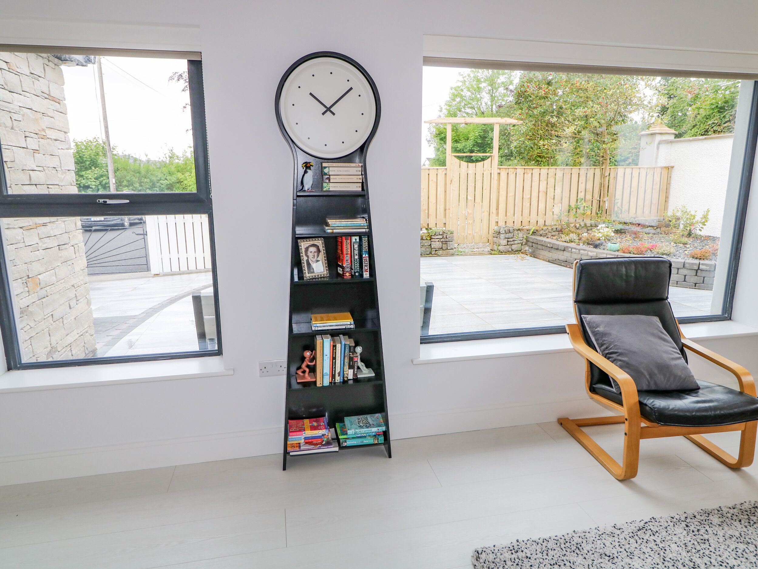 A room with a bookshelf, a clock, a chair, and large windows.