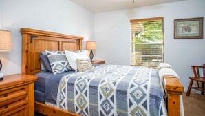 A bedroom with a wooden headboard, a patterned bedspread, a nightstand with a lamp, a window with blinds, and a framed picture on the wall. - Sunnyside | Fire Pit | Spacious & Comfortable (Fredericksburg)