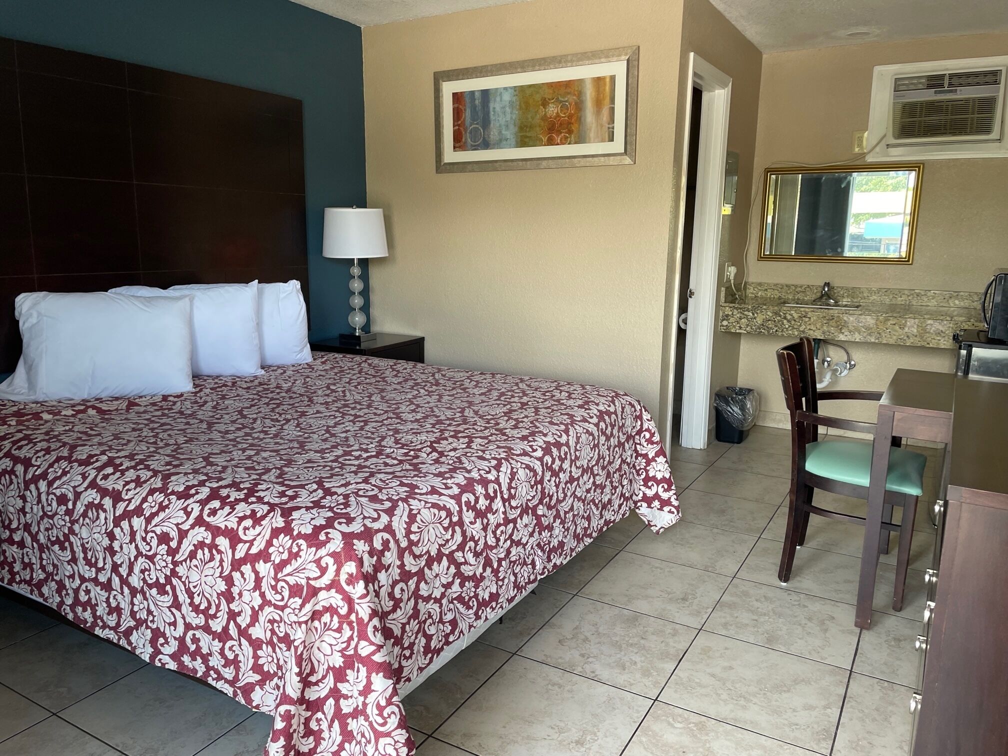 A hotel room with a large bed, a nightstand with a lamp, a bathroom visible through an open door, and a small dining area with a chair and table.