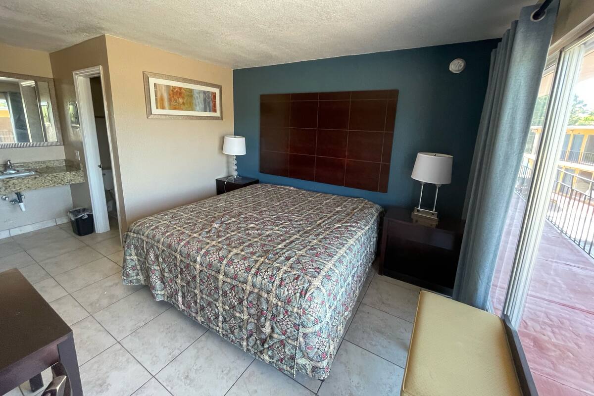 Une chambre d’hôtel comprenant un lit, des tables de chevet, une commode, un miroir, une fenêtre avec des rideaux et une vue sur le balcon extérieur.