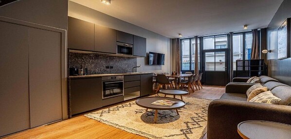 A modern kitchen with dark cabinetry, a built-in oven, and a microwave. There is a dining area with a table and chairs, and a sofa in the living room.