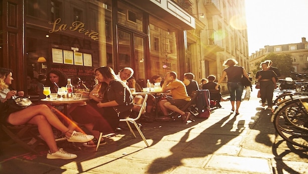Un café al aire libre con mesas y sillas, gente sentada y caminando cerca.