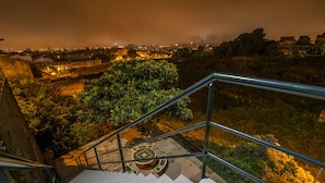 Una escalera con barandillas de metal que sube a un nivel superior, rodeada de vegetación y un paisaje urbano nocturno.