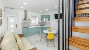 Cocina moderna con armarios azul verdoso, encimera blanca y estufa integrada. Se observa un comedor con una mesa blanca y dos sillas, una amarilla y otra gris. A la derecha, se ve una escalera de madera.