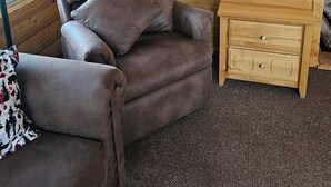 A cozy room with a wooden log wall, a brown armchair with a patterned throw, a small wooden nightstand, and a lamp.