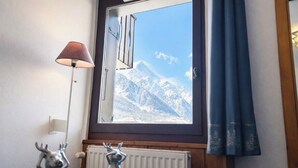 A room with a window overlooking snow-capped mountains, a table with two deer figurines, and a lamp.