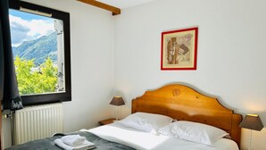 A neatly made bed with white linens and towels, a wooden headboard, two bedside lamps, a framed picture on the wall, and a window with a mountain view.