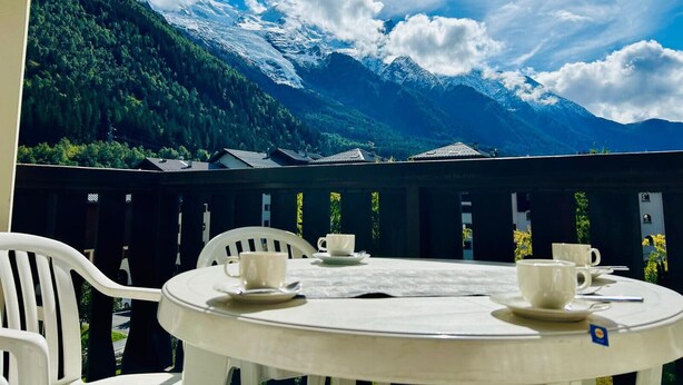 A round table with white chairs and four white cups on a balcony overlooking snow-capped mountains and a forested valley.