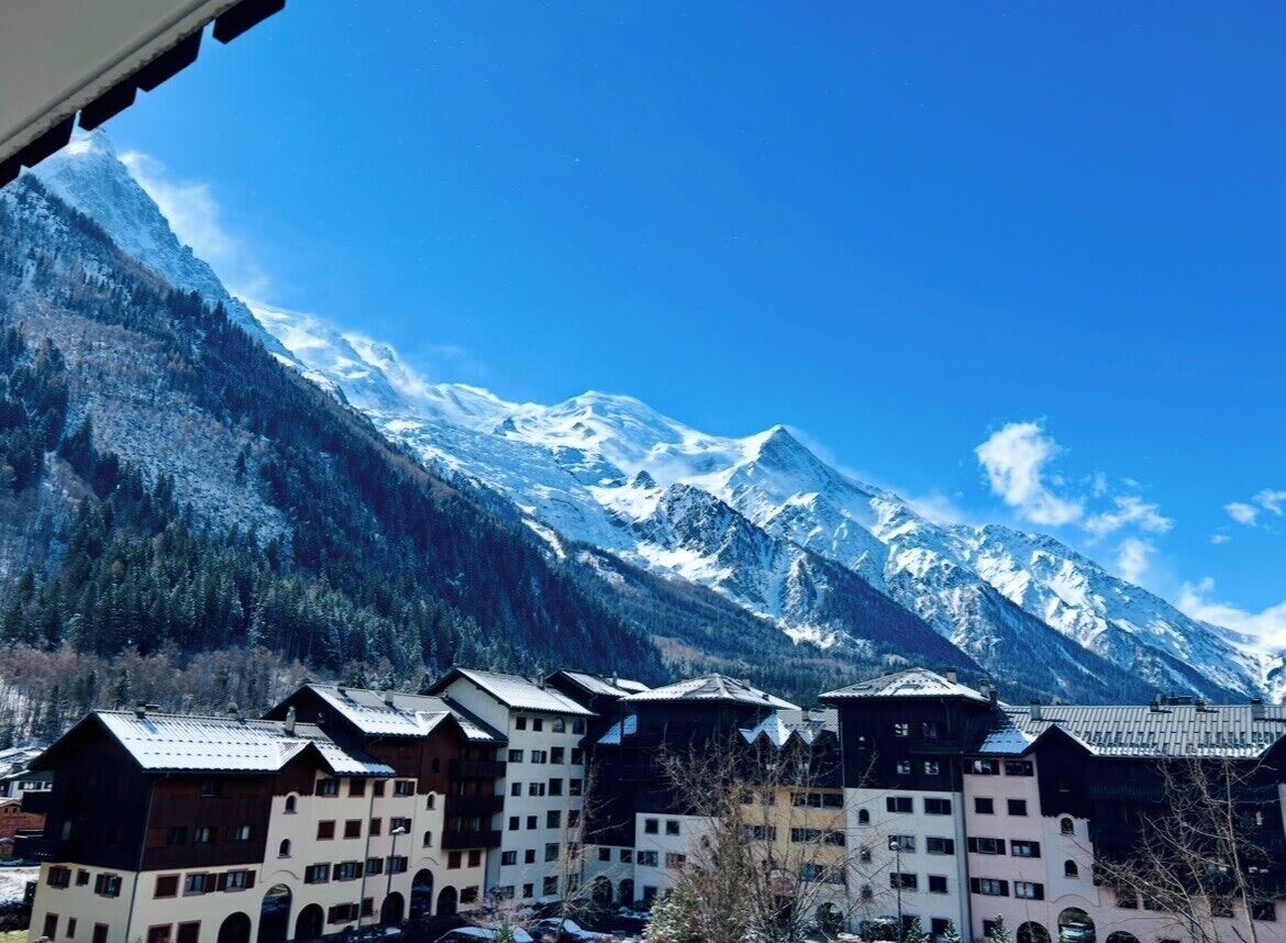 A mountain resort with chalet-style buildings, snow-capped peaks, and clear blue sky.