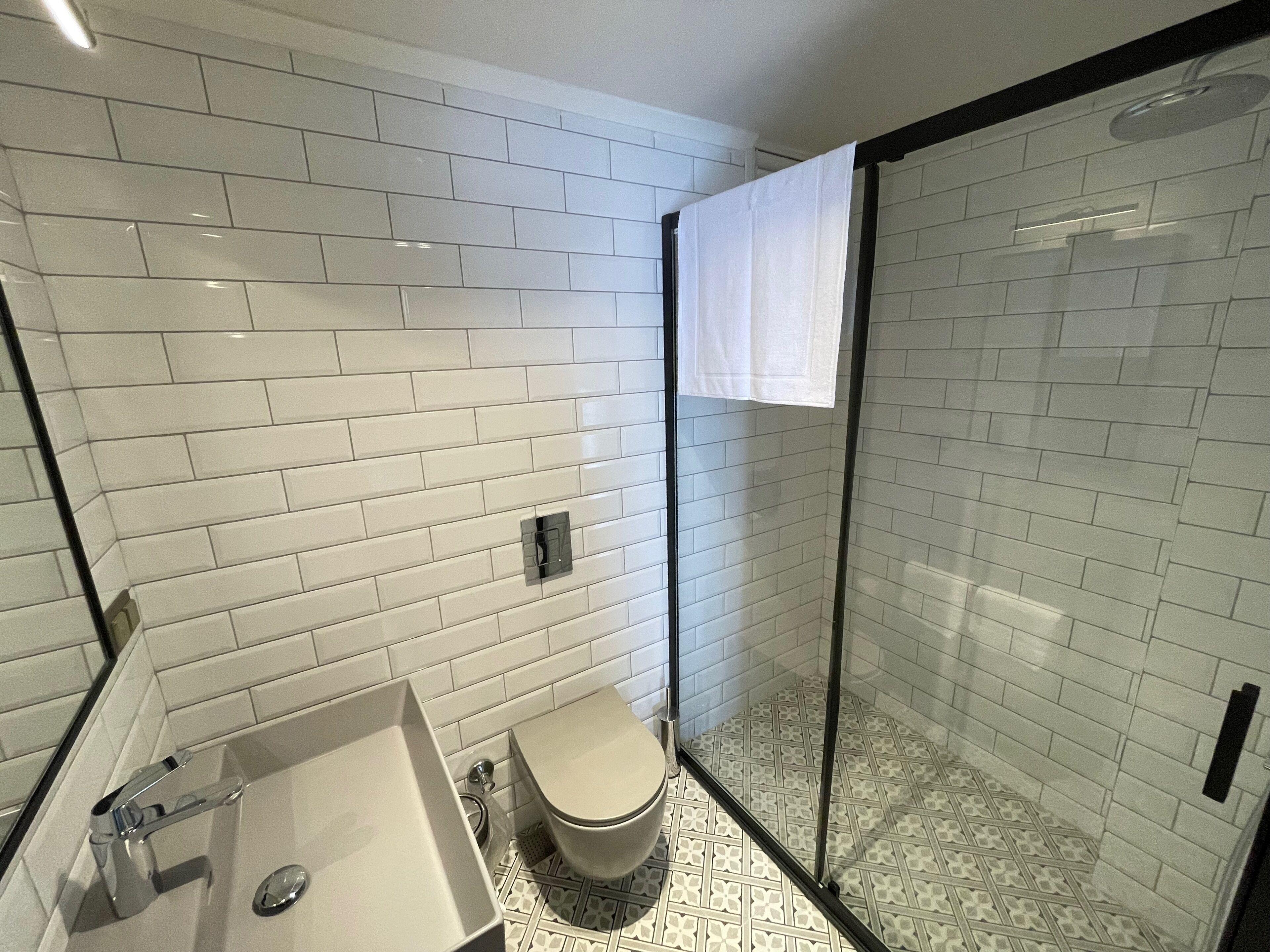 Une salle de bain moderne avec des carreaux de céramique blancs rectangulaires, une douche en verre, un lavabo blanc et des toilettes.