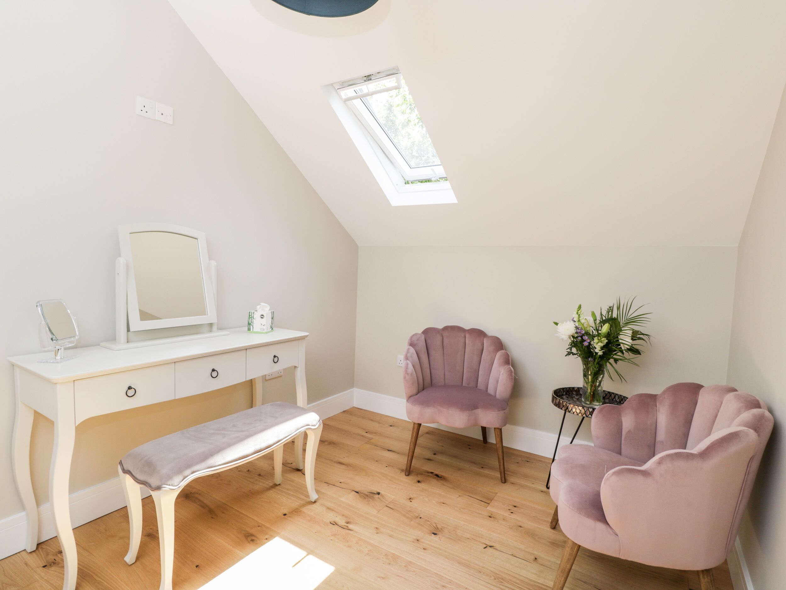 A bedroom with a sloped ceiling, a vanity with a mirror, two pink chairs, a bench, and a potted plant.
