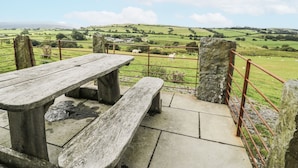 Un espace extérieur rustique aménagé avec une table et des bancs en pierre, offrant une vue sur un paysage verdoyant avec des collines et des moutons.